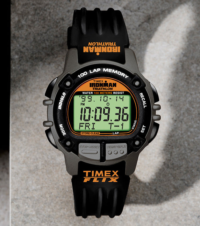 TIMEX® IRONMAN® Flix 100 digital sports watch with a black and gray case, orange accents, and green display, placed on a concrete surface casting a curved shadow.