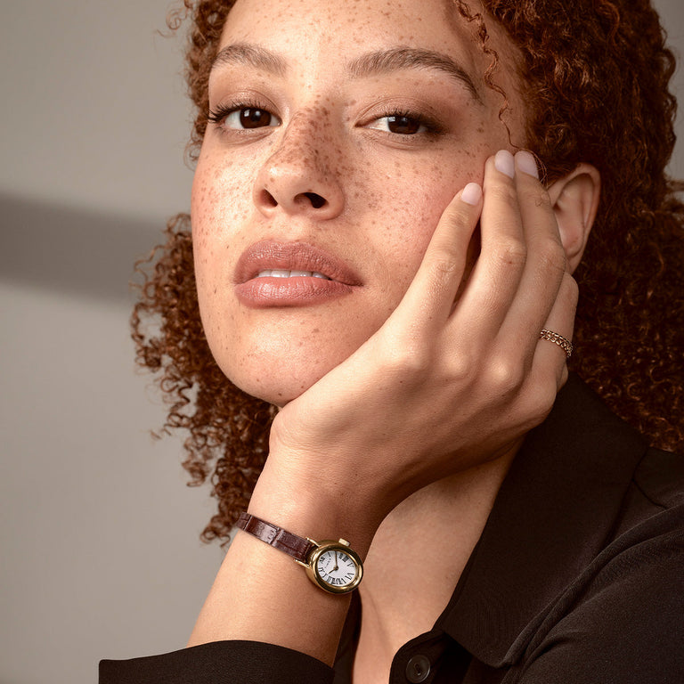 Person wearing the TIMEX® Cavatina watch with a gold-tone case and brown leather strap, paired with a black outfit in a softly lit setting.