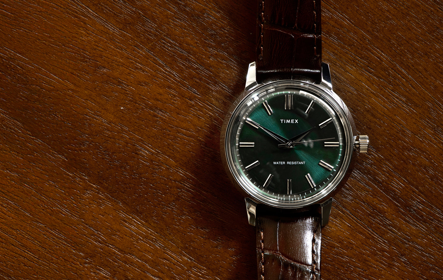 A Timex x Todd Snyder wristwatch with a deep green sunburst dial, silver hour markers, and a brown leather strap, shown resting on a textured wooden surface.