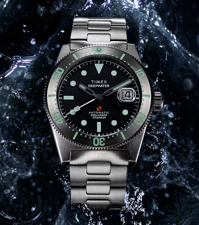 Timex Deepwater Reef 200 Titanium watch with a black dial and aqua lume markers, shown against a dramatic backdrop of splashing water.