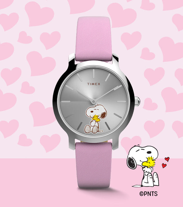 Peanuts® x Timex Transcend Snoopy Hug watch with a silver sunray dial, pink leather strap, and Snoopy hugging Woodstock on the dial, shown against a pink heart background.
