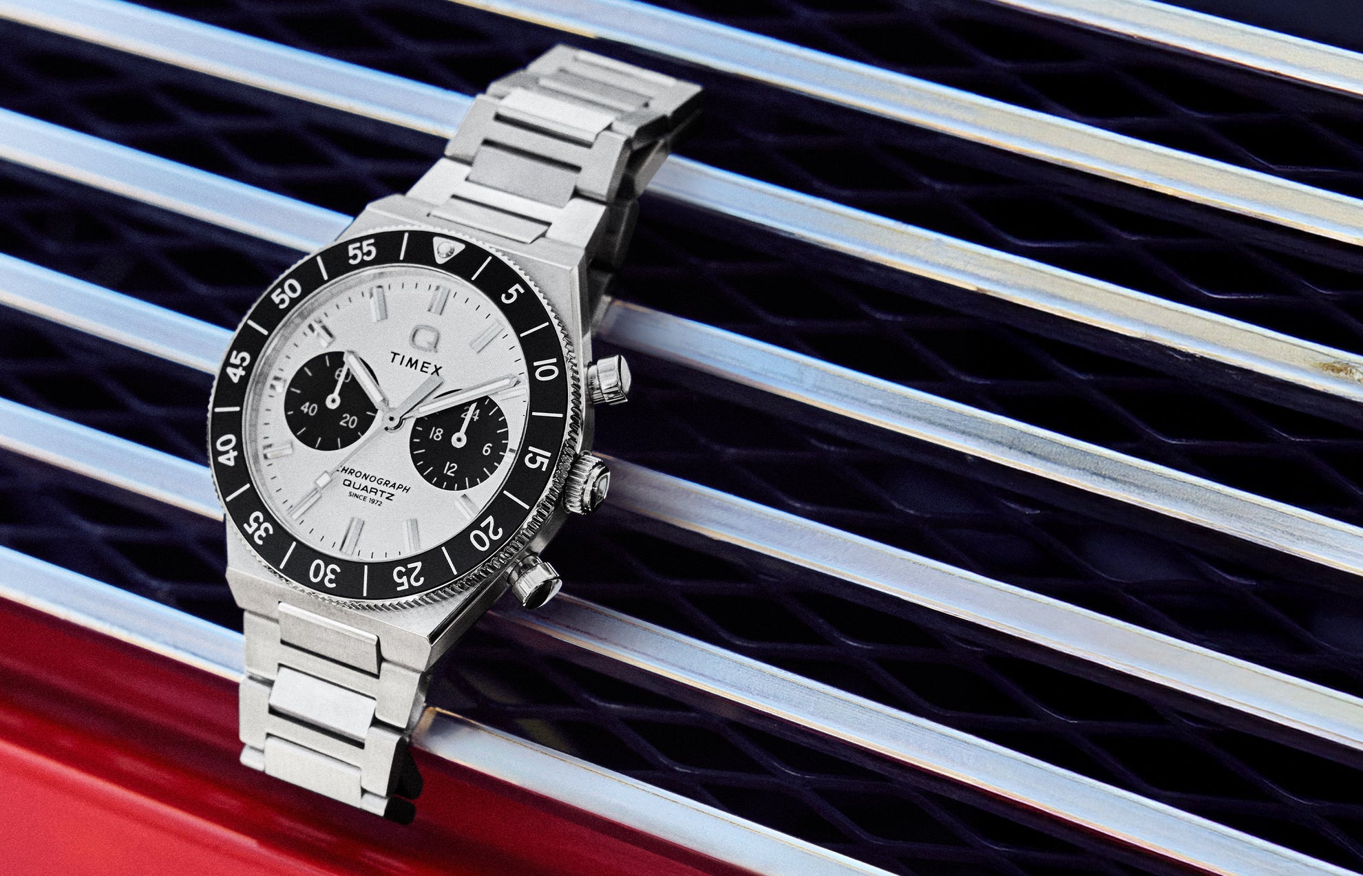 Q Timex Continental Chronograph with silver dial, black bezel, and stainless steel bracelet resting on a metal grille surface.