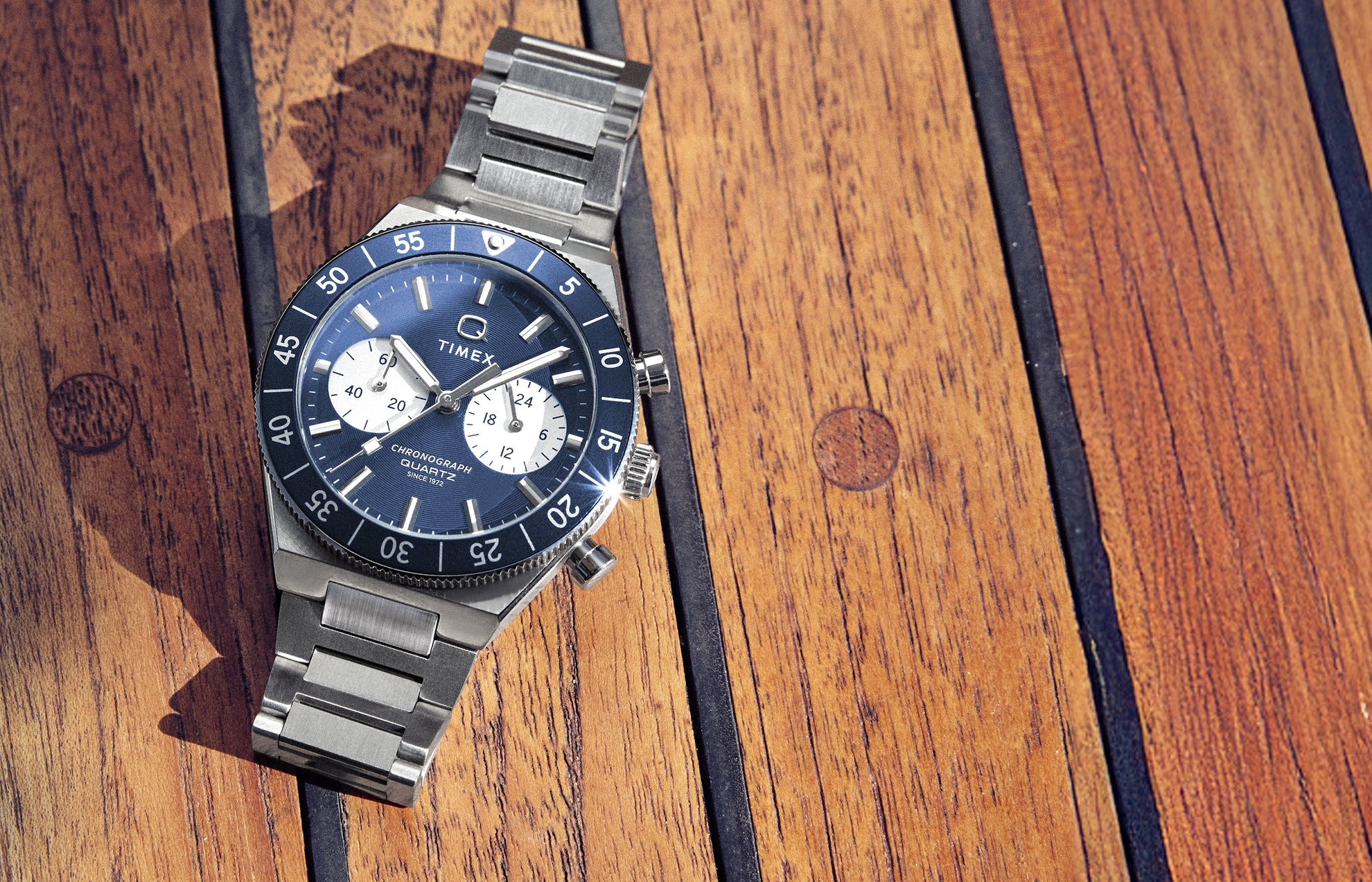Q Timex Continental Chronograph with blue dial, black bezel, and stainless steel bracelet resting on a wooden deck surface.