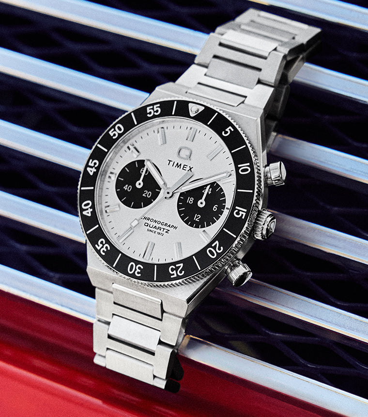 Q Timex Continental Chronograph with silver dial, black bezel, and stainless steel bracelet resting on a metal grille surface.