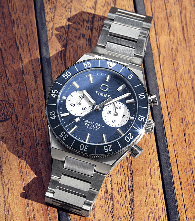 Q Timex Continental Chronograph with blue dial, black bezel, and stainless steel bracelet resting on a wooden deck surface.