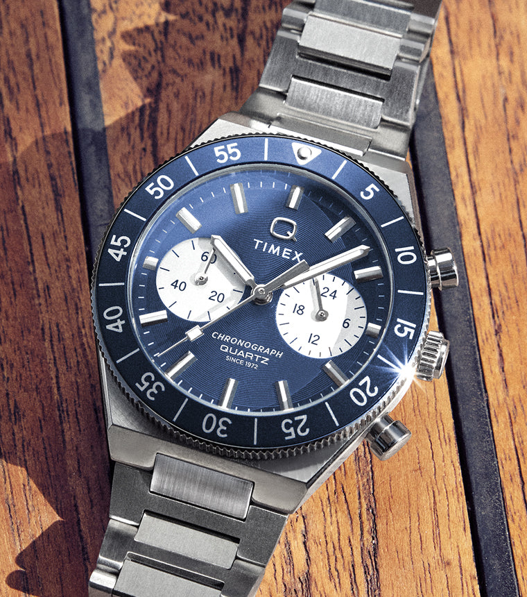 Timex Q Continental Chrono stainless steel watch with a blue dial, white chronograph subdials, and rotating blue bezel, photographed on a wooden surface.