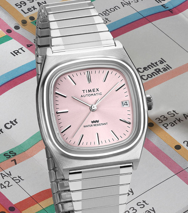 Timex® Automatic 1983 E Line stainless steel watch with light pink sunray dial displayed at an angle on a vintage subway map.