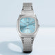 Front view of the Timex® Automatic 1983 E Line watch featuring a light blue dial, silver-tone case, and stainless steel expansion bracelet on a clean background.