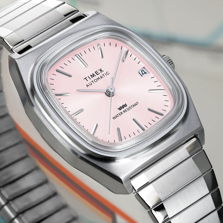 Close-up of the Timex® Automatic 1983 E Line watch showing a light pink sunray dial, applied silver hour markers, date window, and brushed stainless steel case.
