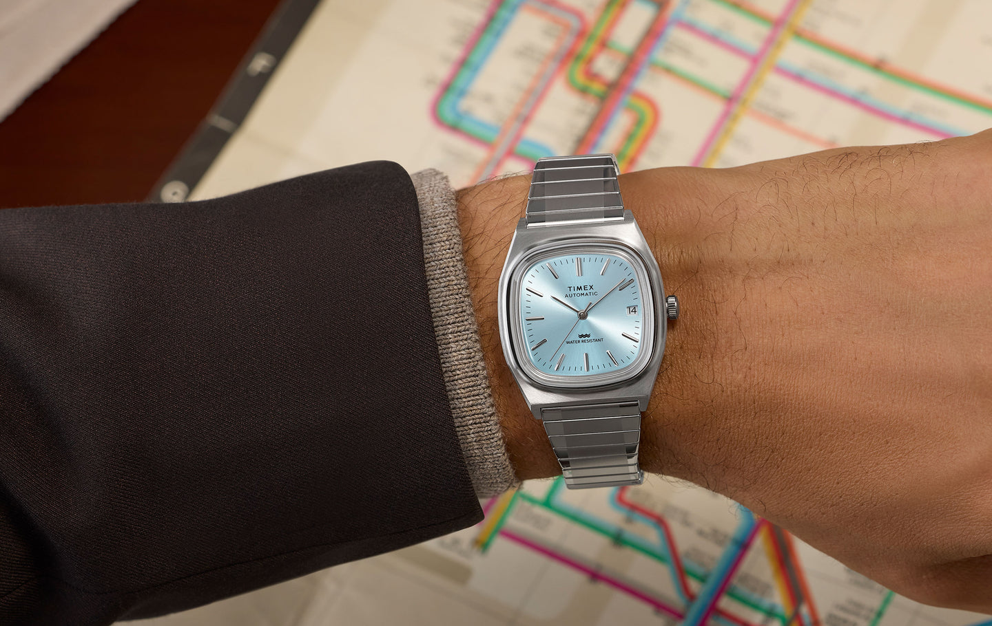 Timex® Automatic 1983 E Line watch with a light blue dial and stainless steel bracelet worn on wrist, featuring a day-date window and brushed silver-tone case, shown against a colorful transit map background.