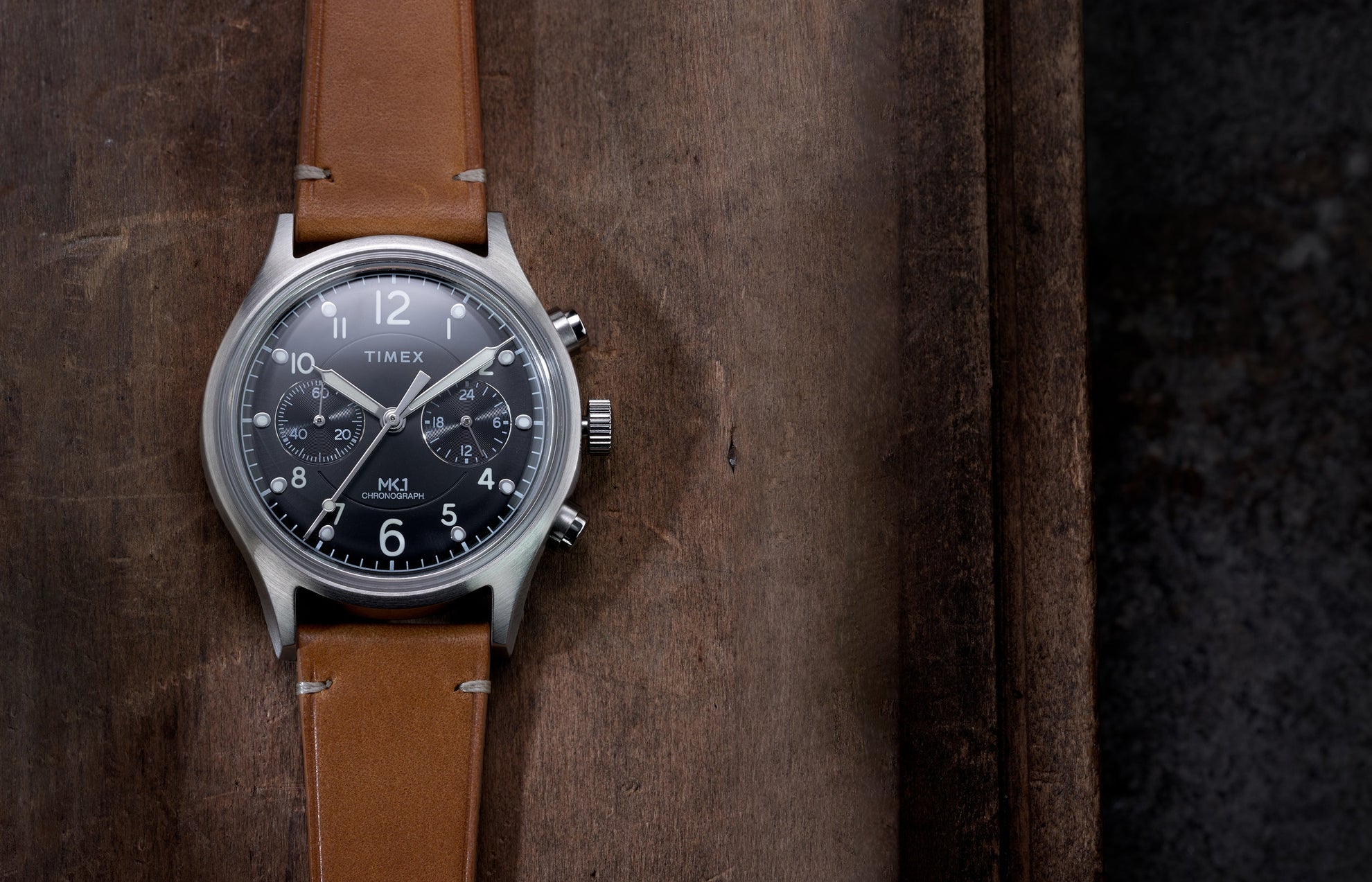 Timex MK1 Chronograph with brown leather strap and black dial, displayed flat on a rustic wooden surface.