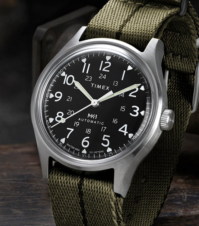 Timex MK1 Automatic with green fabric strap and black dial, displayed upright on a rugged industrial surface.