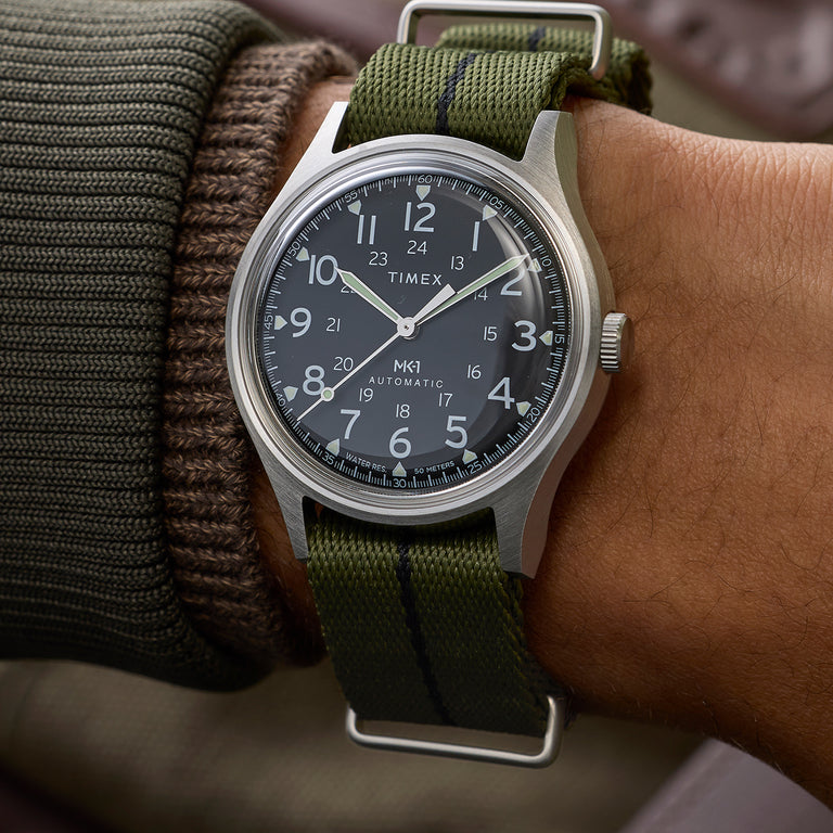 Timex MK1 Automatic watch with a black dial and white Arabic numerals, brushed silver-tone case, and green fabric strap, shown worn on the wrist with a casual knit sleeve.