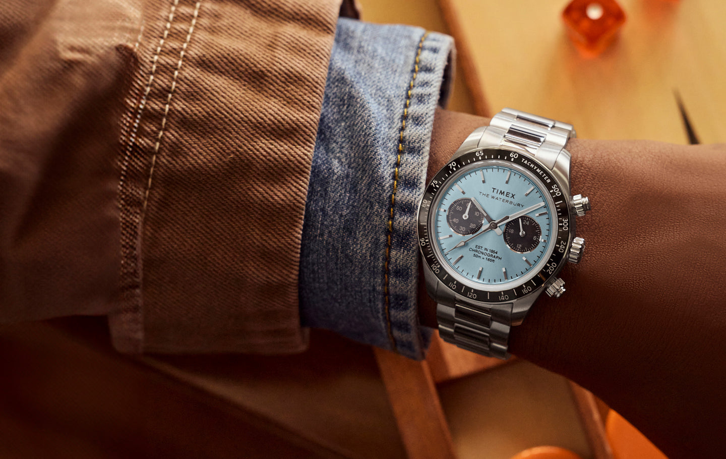 Close-up of a person’s wrist wearing a Timex Waterbury Heritage chronograph watch with a light blue dial, black tachymeter bezel, and stainless steel bracelet. The watch features two black subdials and silver-tone hour markers, shown against a warm-toned background with a brown jacket and denim shirt cuff.