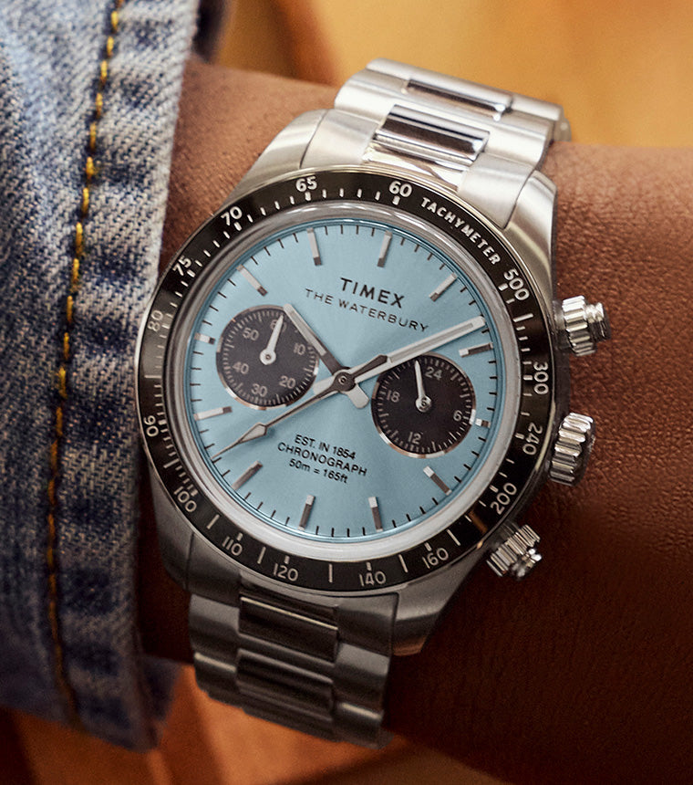 Close-up of a person’s wrist wearing a Timex Waterbury Heritage chronograph watch with a light blue dial, black tachymeter bezel, and stainless steel bracelet. The watch features two black subdials and silver-tone hour markers, shown against a warm-toned background with a brown jacket and denim shirt cuff.