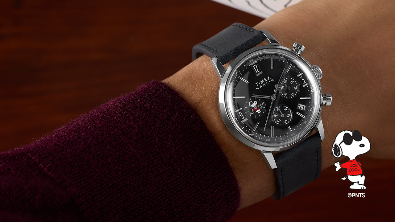 Peanuts® x Timex Marlin® Chronograph Joe Cool watch worn on a wrist, featuring a stainless steel case, black leather strap, black chronograph dial, and Snoopy as Joe Cool in a red shirt on a subdial.