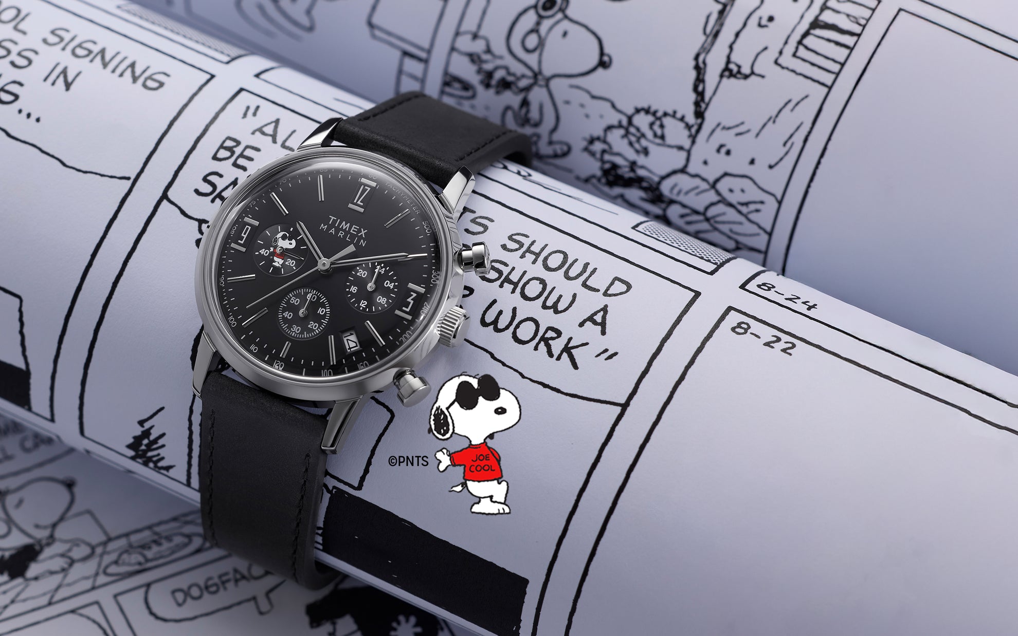 Peanuts® x Timex Marlin® Chronograph Joe Cool watch with a stainless steel case and black leather strap, displayed on Peanuts comic strip pages, showing a black chronograph dial with Snoopy Joe Cool artwork.