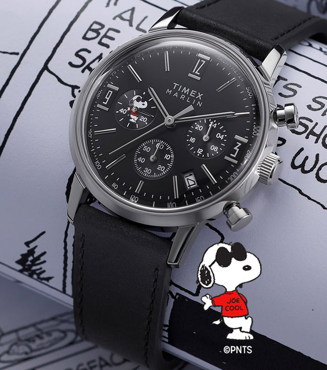 Peanuts® x Timex Marlin® Chronograph Joe Cool watch with a stainless steel case and black leather strap, displayed on Peanuts comic strip pages, showing a black chronograph dial with Snoopy Joe Cool artwork.