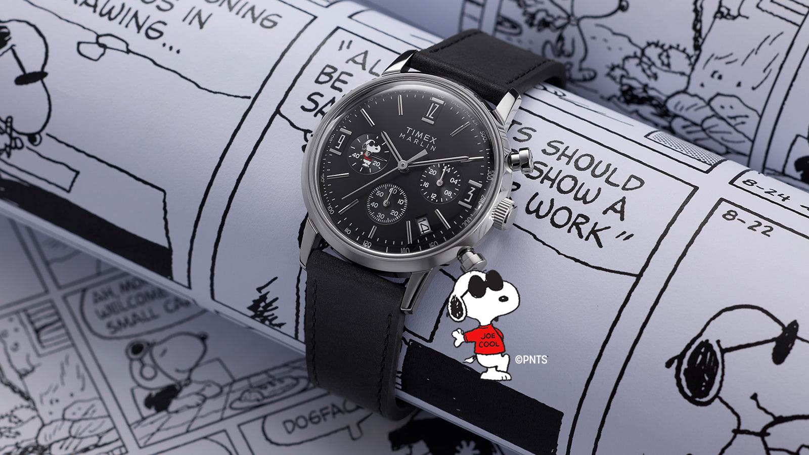 Peanuts® x Timex Marlin® Chronograph Joe Cool watch resting on illustrated Peanuts comic pages, featuring a black dial with silver markers, chronograph subdials, and Snoopy Joe Cool detail.