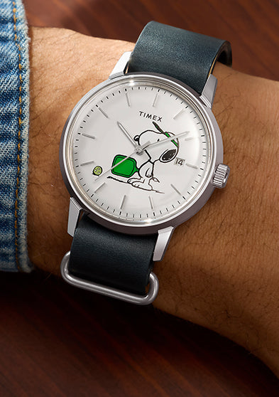 Peanuts® x Timex Marlin® Automatic Pickleball watch worn on wrist, featuring Snoopy playing pickleball on a white dial, stainless-steel case, date window, and dark green leather strap.