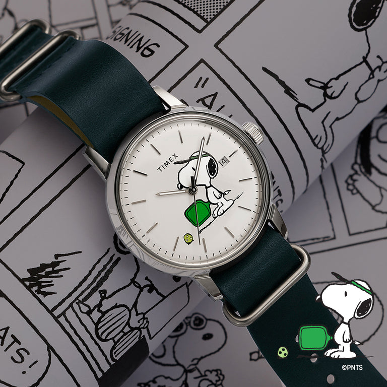 Peanuts® x Timex watch with a silver case, white dial featuring Snoopy holding a green pickleball paddle, green fabric strap, and date window, styled on top of a Peanuts comic strip page.