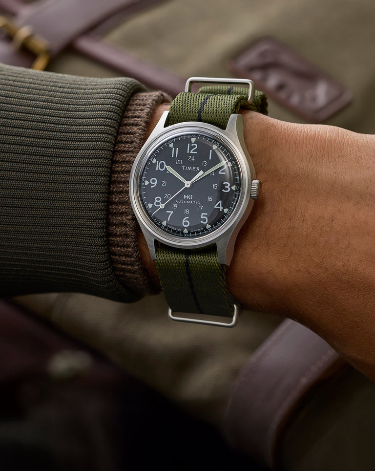 Timex MK1 Automatic watch with a black dial and green fabric strap worn on wrist, featuring white Arabic numerals and luminous hands.