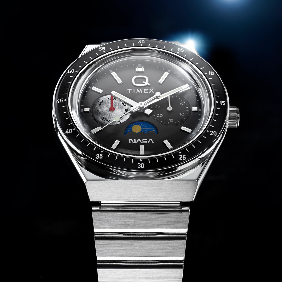 Close-up of Q Timex® x NASA watch featuring brushed stainless steel bracelet, black bezel, gray dial with day subdial, chronograph-style subdial, and NASA logo at 6 o’clock.