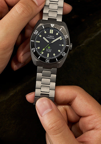 Expedition® Pioneer Titanium Automatic watch held in hand, featuring a brushed titanium case and bracelet, black dial, and green seconds hand.