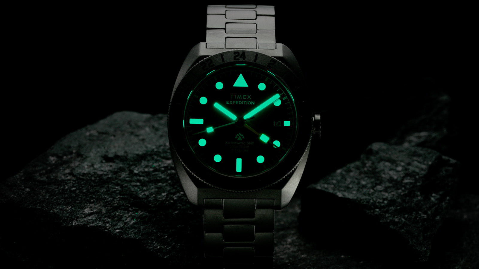 Low-light image of the Expedition® Pioneer Titanium Automatic GMT with fully illuminated hands and hour markers glowing green against the dark dial.