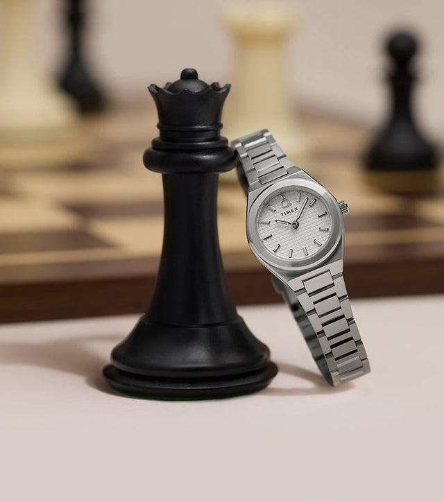 Close-up of Q Timex® Continental Mini stainless steel watch with silver textured dial posed beside a black chess piece.