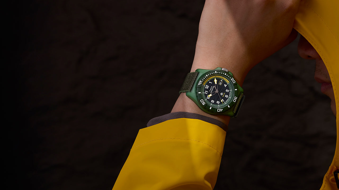 Timex Expedition® Freedive Solar watch with green case and black dial worn on wrist with yellow rain jacket, showing black fabric strap and bold white markers.