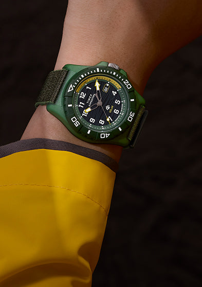 Timex Expedition® Freedive Solar watch with green case and black dial worn on wrist with yellow rain jacket, showing black fabric strap and bold white markers.
