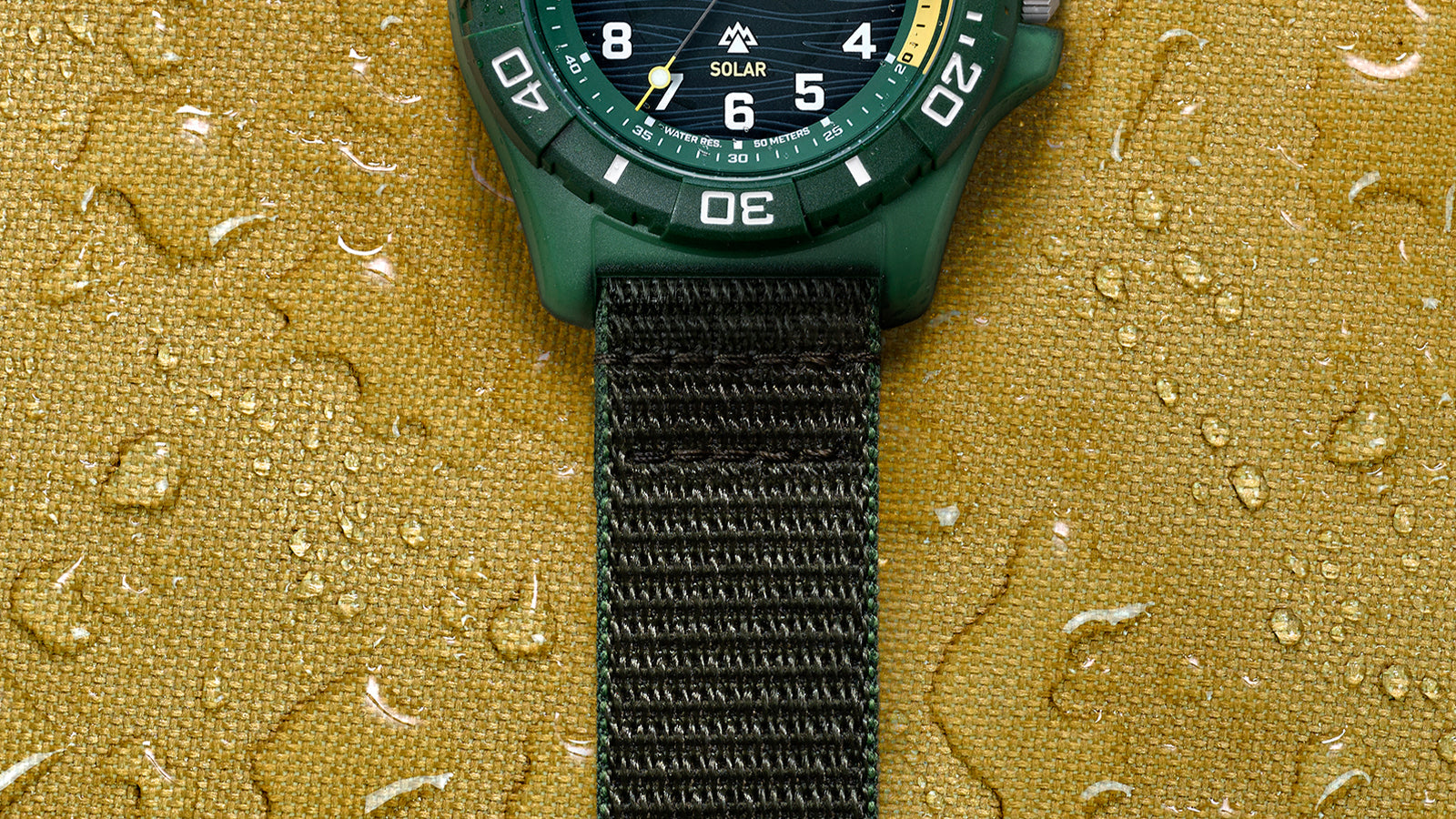 Detail of black fabric strap and green case of Timex Expedition® Freedive Solar watch on yellow water-resistant background with droplets.