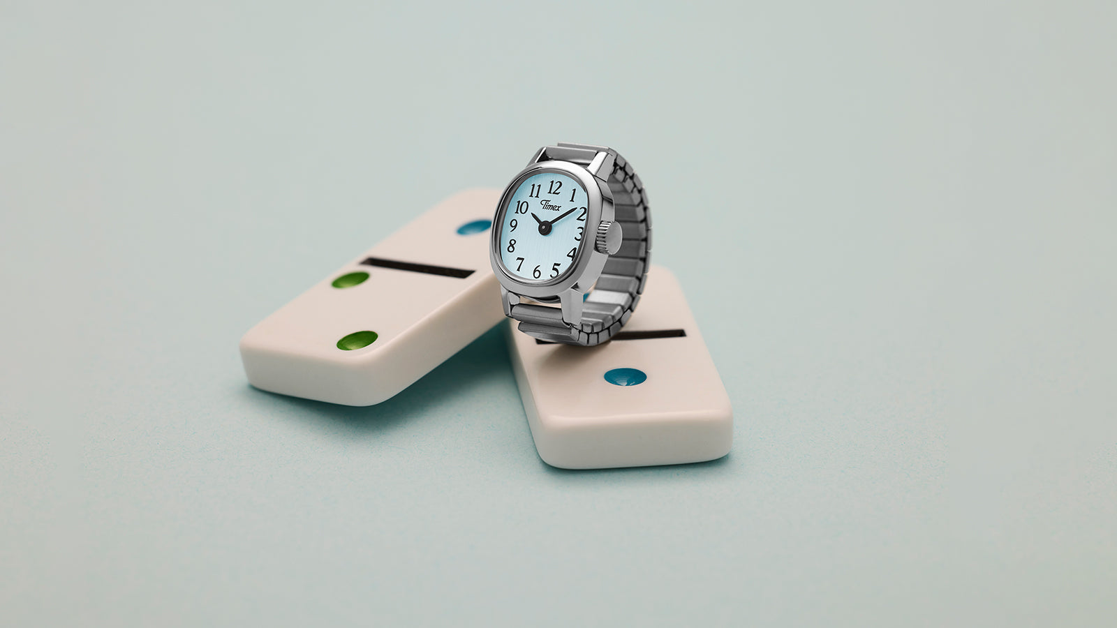 Timex Cavatina® Mini rectangular watch with silver-tone expansion bracelet styled on domino props against pastel blue backdrop.