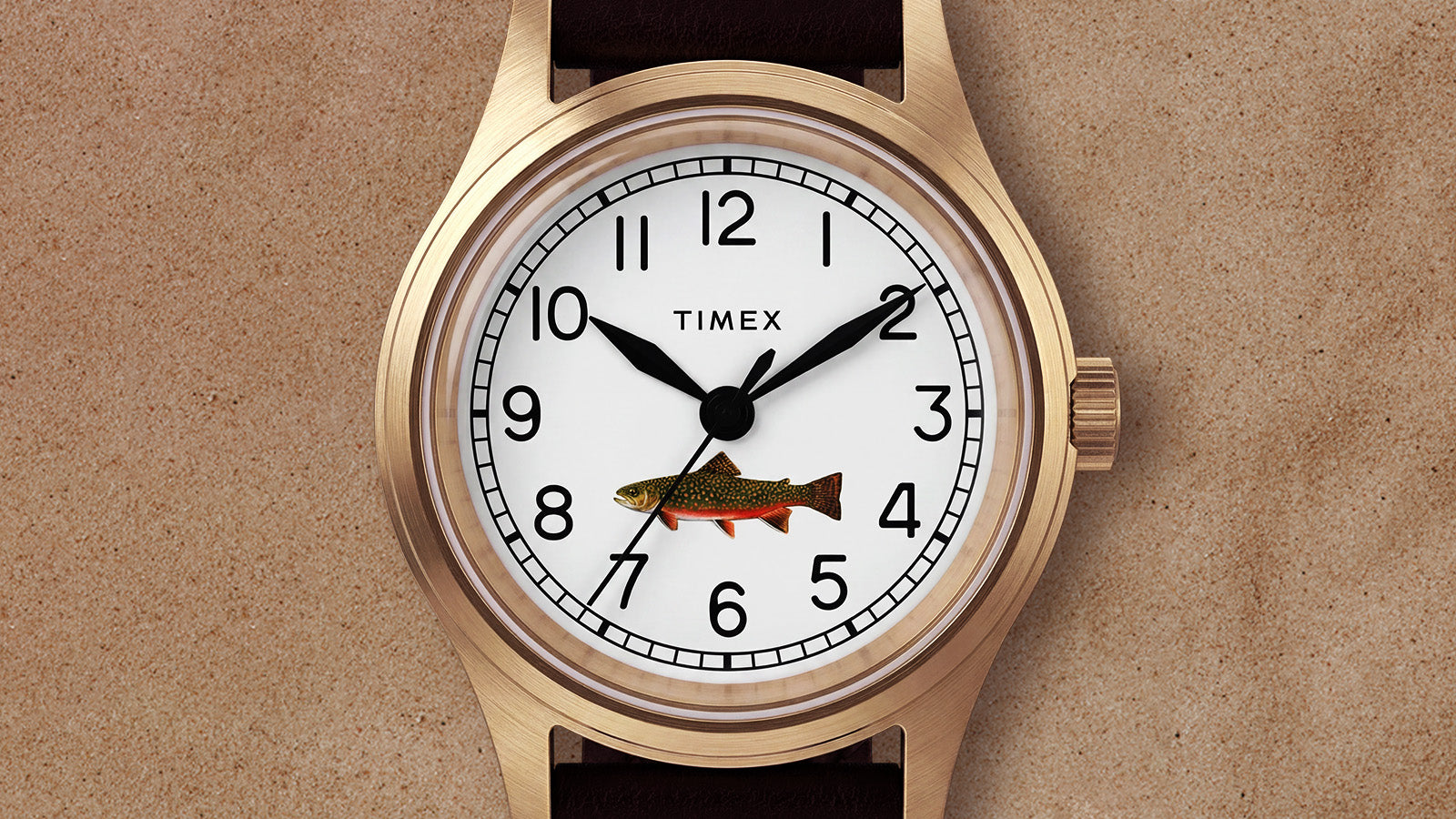 Close-up of Timex MK1 watch face with white dial, black numerals, and colorful fish graphic on a gold-tone case against a tan background.
