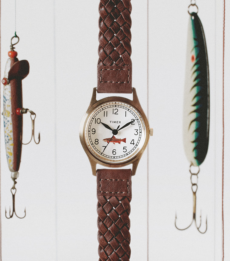 Timex MK1 watch with braided brown leather strap suspended among colorful vintage fishing lures against a soft white background.