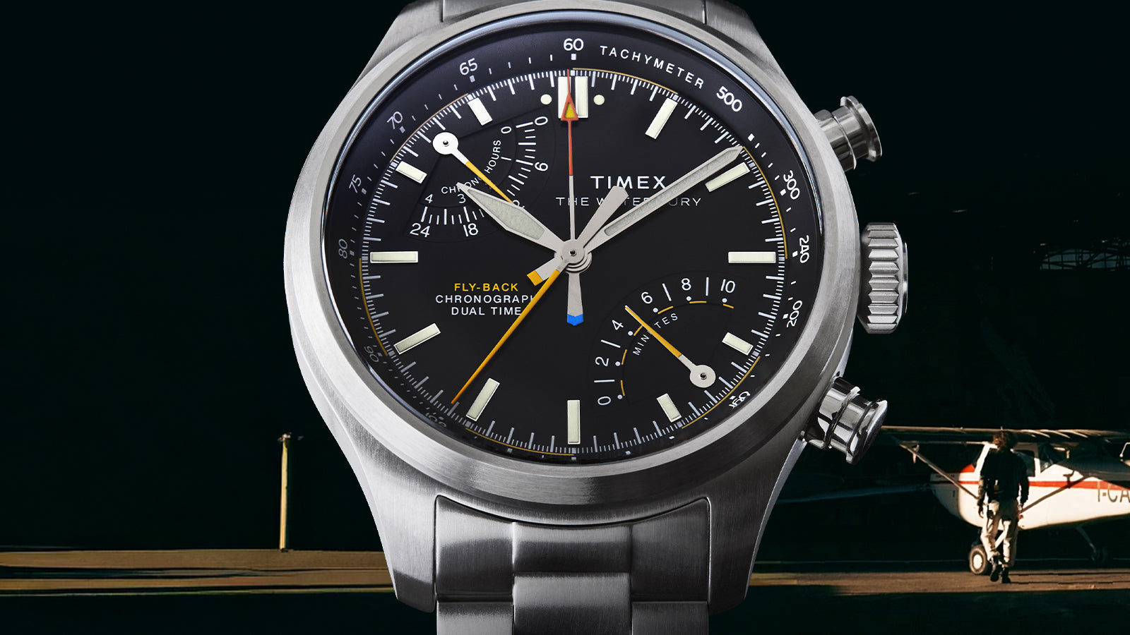 Timex Waterbury Ace Fly-Back Chronograph pilot watch with stainless steel bracelet against aircraft hangar background