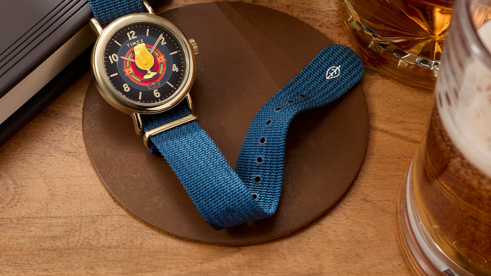 Harry Potter x Timex Weekender Butterbeer watch with gold-tone case and blue fabric strap displayed beside books and a Butterbeer-inspired drink.