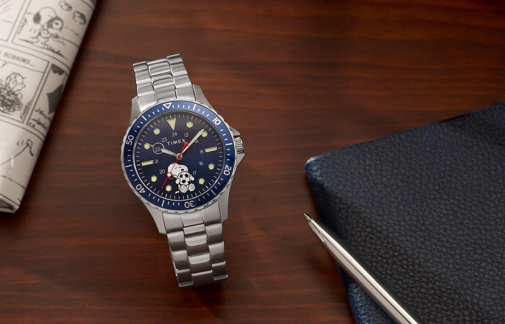 Timex Navi x Peanuts Snoopy soccer watch with blue dial and stainless steel bracelet on wooden desk with notebook and pen