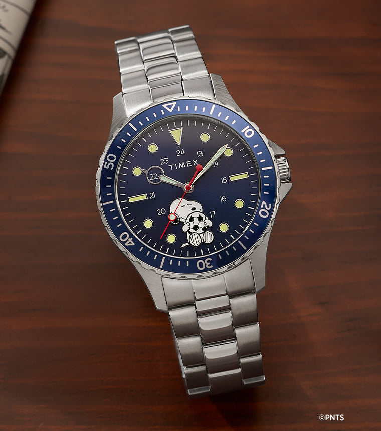 Timex Navi x Peanuts Snoopy soccer watch with blue dial and stainless steel bracelet on wooden desk with notebook and pen