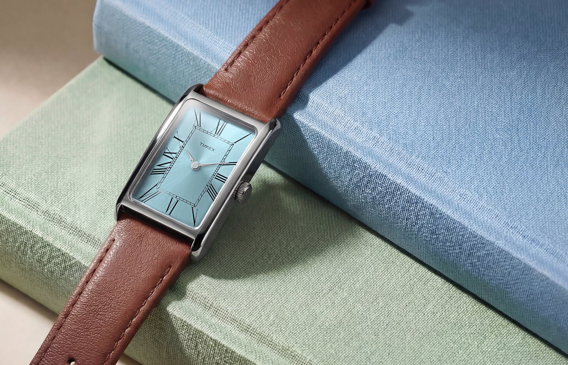 Timex® 1976 Lexington Reissue Inspired watch with a light blue sunray dial, Roman numeral markers, rectangular case, and brown leather strap displayed on stacked books.