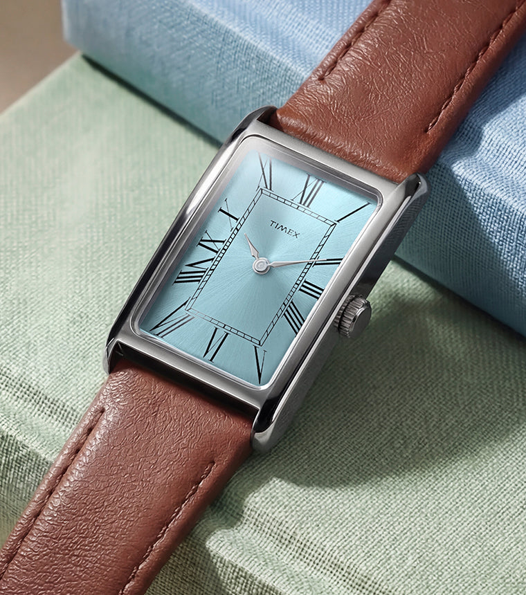 Timex® 1976 Lexington Reissue Inspired watch with a light blue sunray dial, Roman numeral markers, rectangular case, and brown leather strap displayed on stacked books.