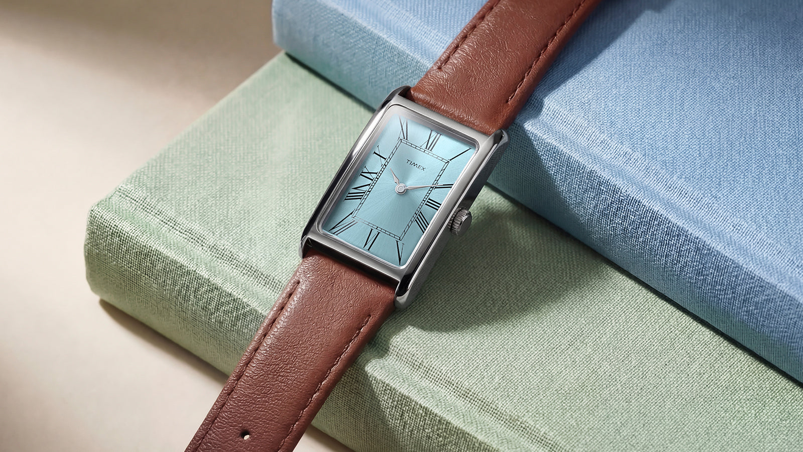 Angled view of the Timex® 1976 Lexington Reissue Inspired watch resting on books, featuring a light blue dial with Roman numerals and a brown leather strap.