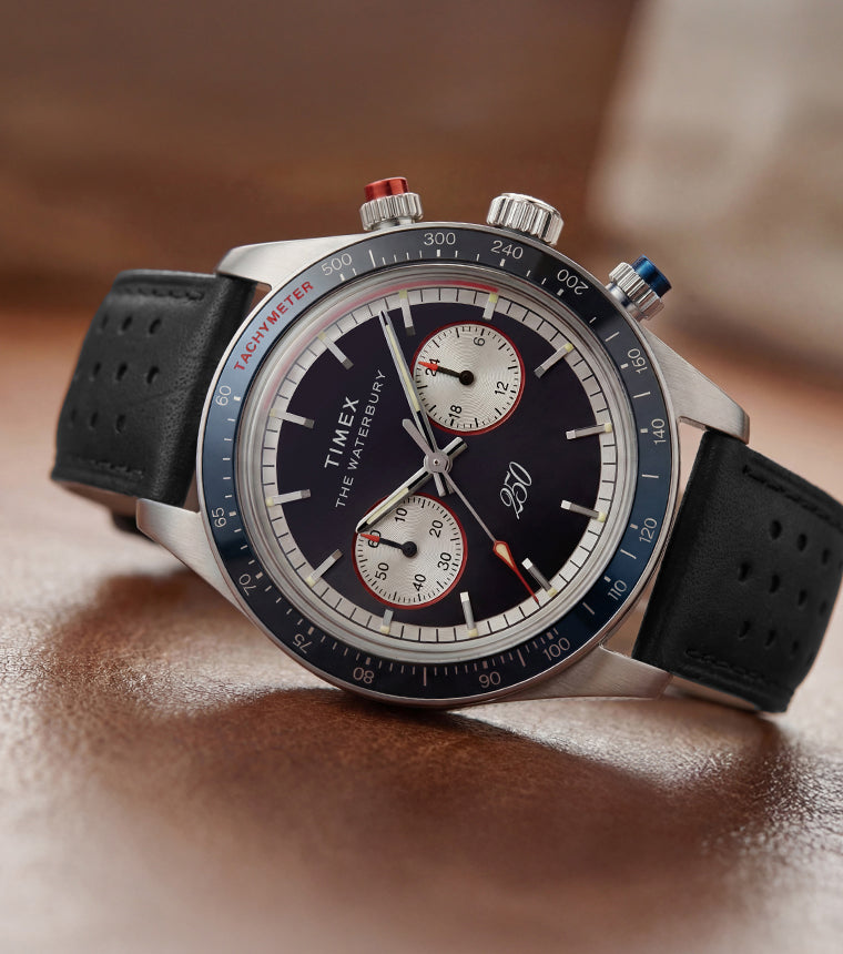 Timex Waterbury Heritage America 250 chronograph watch with a navy dial, stainless steel case, black perforated leather strap, and tachymeter bezel on a brown leather surface.