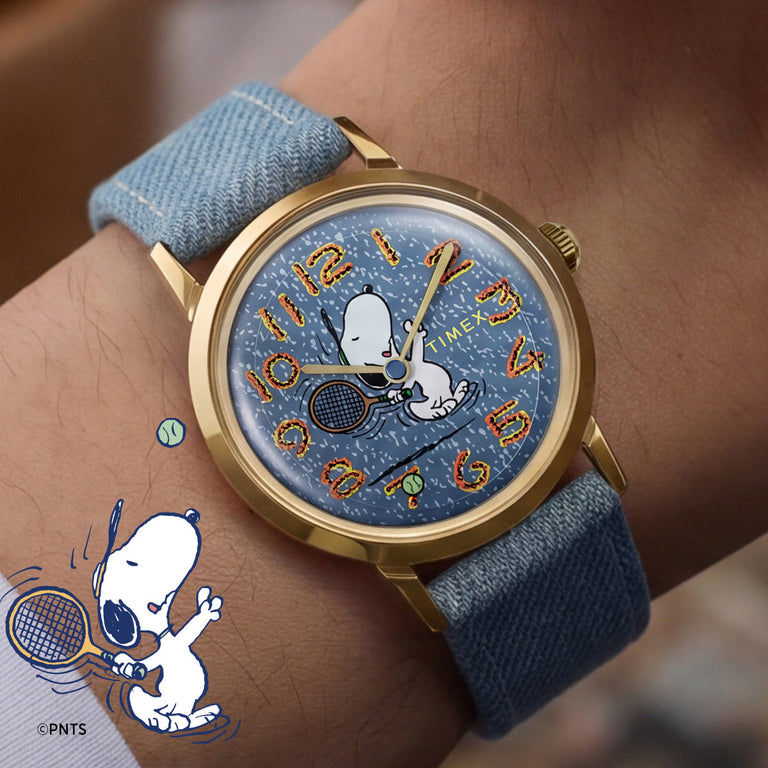 Close-up of a Peanuts® x Timex Marlin® watch on a wrist, featuring Snoopy playing tennis on a blue textured dial. The watch has a gold-tone case, denim-blue fabric strap, and colorful stylized numerals, blending playful Peanuts artwork with a vintage-inspired design.