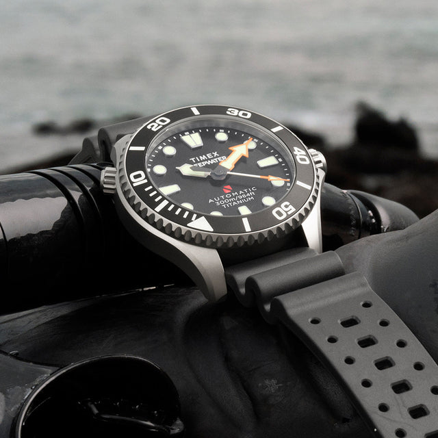 Timex Deepwater Meridian 300 Titanium HEV Automatic dive watch resting on scuba diving gear near the ocean shoreline.