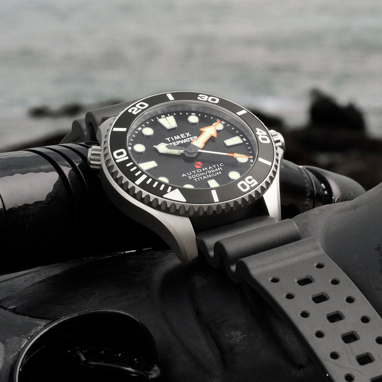 Timex Deepwater Meridian 300 Titanium HEV Automatic dive watch resting on scuba diving gear near the ocean shoreline.