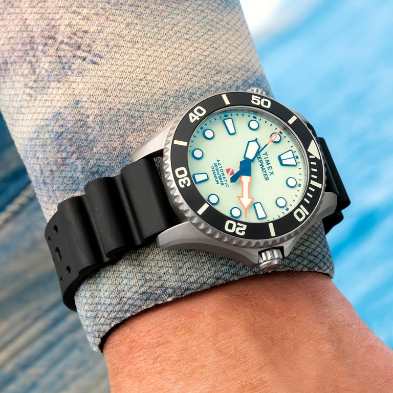 Lifestyle image of the Timex Deepwater Meridian 300 Titanium HEV Automatic watch worn on the wrist, featuring a luminous white dial with bold blue hands and markers, an orange seconds hand, a black unidirectional dive bezel, a titanium case, and a black rubber strap against a blue waterside background.