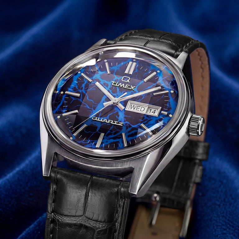 Close-up product image of a Q Timex watch with a silver-tone stainless steel case and black leather strap. The dial features a striking blue lightning-inspired Vega pattern with silver hour markers and hands, plus a day-date window displaying ‘WED 14.’ The watch is photographed against a rich blue satin fabric background, emphasizing its bold electric-blue design and polished finish.