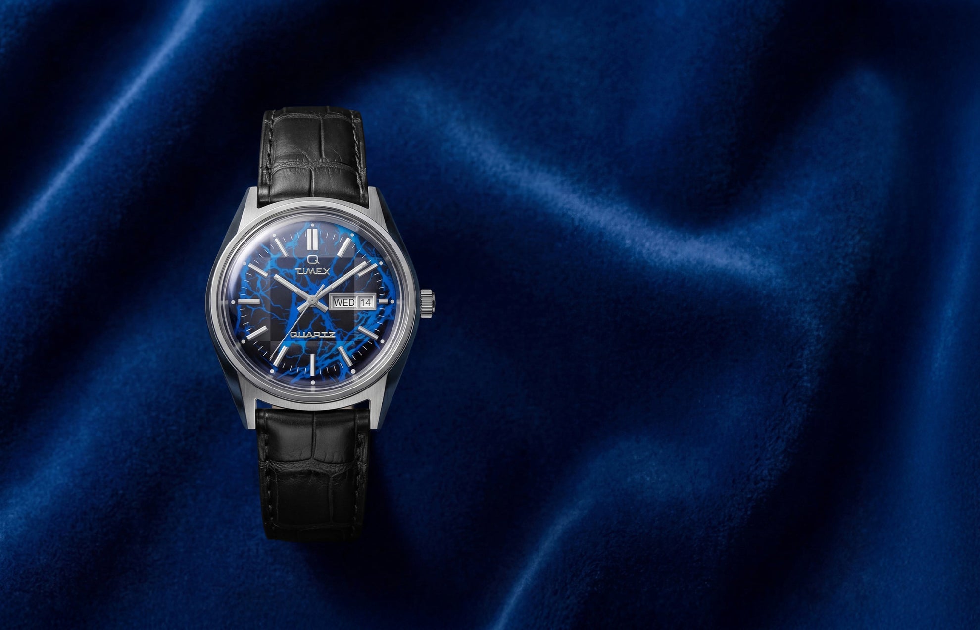 Q Timex Blue Vega watch with electric blue lightning-pattern dial on black leather strap against deep blue satin background